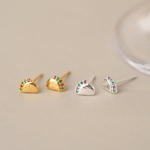 May include: Two pairs of taco-shaped stud earrings, one pair in gold-tone metal and one pair in silver-tone metal. Each earring features small, embedded green and pink stones along the curved edge and tiny dot indentations on the surface.