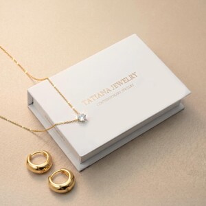 May include: A gold chain necklace with a single clear round stone pendant and a pair of matching gold hoop earrings. The jewelry is displayed with a white rectangular box featuring the gold text TATIANA JEWELRY CONTEMPORARY JEWELRY.