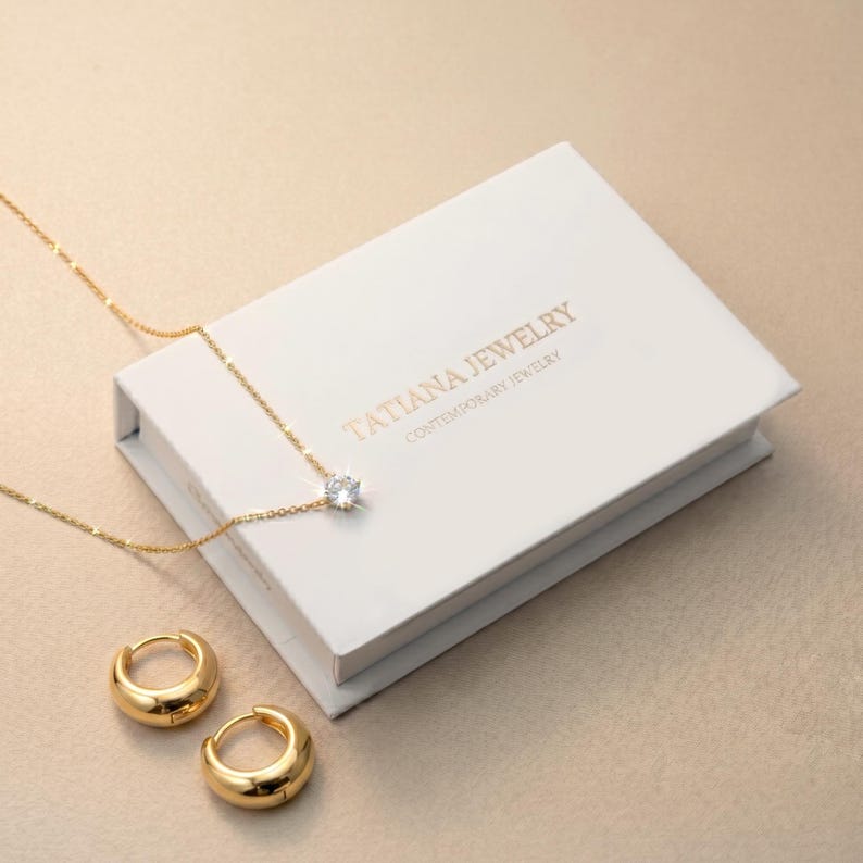 May include: A gold chain necklace with a single clear round stone pendant and a pair of matching gold hoop earrings. The jewelry is displayed with a white rectangular box featuring the gold text TATIANA JEWELRY CONTEMPORARY JEWELRY.