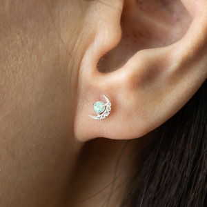 May include: A silver stud earring in the shape of a crescent moon with a central opal-like stone and small, clear crystals. The earring is worn on an ear, showcasing its delicate design and sparkle. The earring is a piece of jewelry.