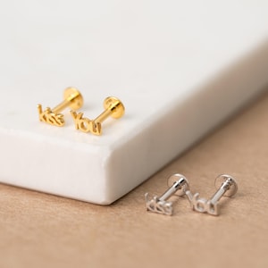May include: Two pairs of flat back stud earrings, one gold-toned and one silver-toned. Each earring features the word kiss on one stud and the word you on the other, written in a cursive script font. The studs have a post and a circular back.