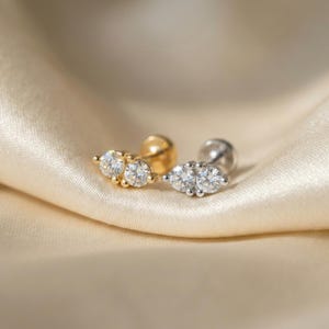 May include: Two stud earrings with three clear, round-cut gemstones. One earring is gold-toned, the other is silver-toned. The earrings are displayed on a cream-colored, satin-like fabric.