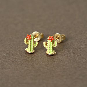 May include: A pair of gold-colored stud earrings featuring a cactus design. Each earring has a light green body with red flower accents. The earrings are set against a brown background.