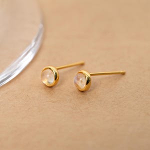 Puede incluir: Un par de pendientes de oro con una piedra preciosa redonda e iridiscente engastada en un bisel. Los pendientes se muestran sobre una superficie neutra de color beige, con un objeto de vidrio curvado y borroso en el fondo.