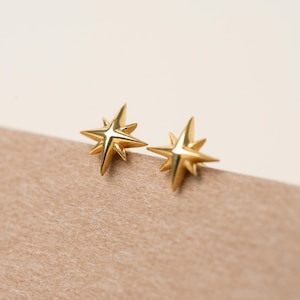 May include: A pair of gold-colored star-shaped stud earrings. Each earring has eight points and a shiny, reflective surface. The earrings are displayed on a brown surface against a neutral background.