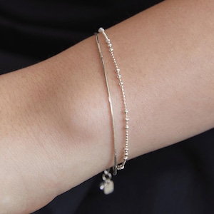Puede incluir: Un delicado conjunto de pulseras de plata. Una es una cadena fina y plana, y la otra presenta pequeñas cuentas espaciadas uniformemente. Ambas pulseras se usan juntas. Un pequeño dije de corazón cuelga del cierre.