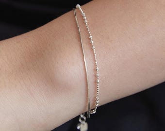 May include: A set of two silver-toned bracelets worn together. One bracelet features a smooth, thin snake chain design, while the other consists of a delicate chain with small, evenly spaced round beads. A small metal charm hangs from the clasp area.