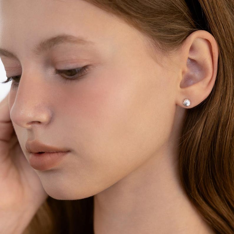 May include: A small, silver-toned stud earring shaped like a seashell. The earring features a textured, fan-like design with vertical ridges, securely fastened to the earlobe. The piece has a polished, metallic finish that reflects light.