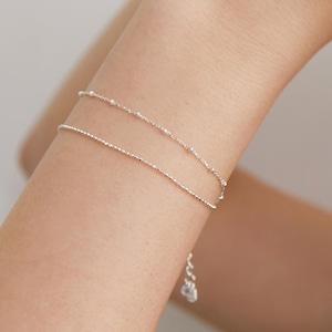 May include: A delicate silver bracelet with two strands. The top strand features small, evenly spaced beads, while the bottom strand has a textured, linear design. The bracelet is shown on a person's arm.