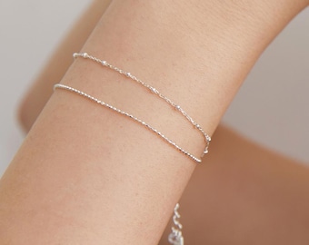 May include: A double-strand silver bracelet featuring one chain with small, evenly spaced spherical beads and a second chain with a textured, shimmering finish. The piece includes a small adjustable extender chain with a tiny charm at the end.