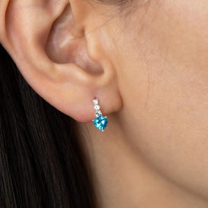 Pode incluir: Um brinco de prata com uma pedra preciosa azul brilhante em forma de coração e três pedras preciosas claras menores. O brinco é usado em uma orelha, com foco na joia.