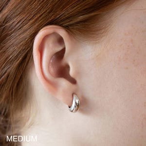 May include: A close-up of a silver hoop earring on an ear. The earring is a medium size and has a smooth, reflective surface. The word "MEDIUM" is printed in white at the bottom of the image.