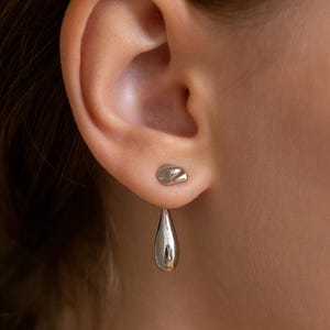 May include: A pair of silver earrings. One earring is a small, rounded stud. The other earring is a teardrop shape that hangs below the earlobe. The earrings are smooth and shiny, reflecting the light.
