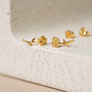 May include: Gold-colored rose earrings. The earrings feature a detailed rose design with a stem and leaves. The earrings are displayed on a light-colored, textured surface. The image shows multiple pairs of earrings.