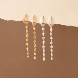 May include: Four pairs of hoop earrings. Two pairs are gold-toned, and two are silver-toned. Each earring features a hoop with small, clear stones and a dangling chain of similar stones. The earrings are displayed against a neutral background.