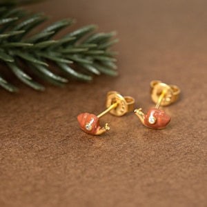 May include: A pair of gold stud earrings shaped like snails with a glossy terracotta-colored enamel shell and a small, clear, round-cut gemstone accent on the body. The earrings feature a standard post back and are crafted with a polished gold finish.