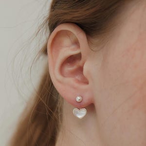 May include: A double-sided earring featuring a small white spherical pearl stud at the front of the earlobe and a heart-shaped charm with a white iridescent inlay framed in silver metal hanging from the back of the earlobe post.