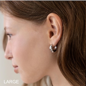 May include: A close-up of a person's ear wearing a silver hoop earring. The earring is a thick, rounded design. The word "LARGE" is printed in white at the bottom left corner of the image.