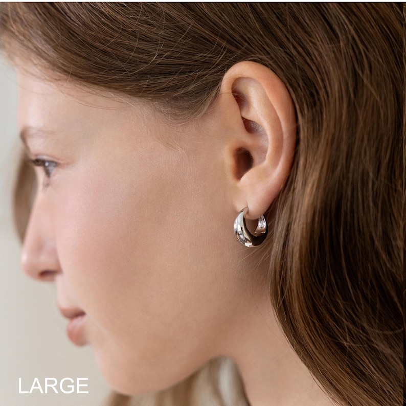 May include: A close-up of a person's ear wearing a silver hoop earring. The earring is a thick, rounded design. The word "LARGE" is printed in white at the bottom left corner of the image.