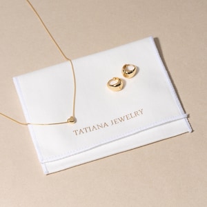 May include: A gold necklace with a heart pendant and two gold hoop earrings are displayed on a white cloth. The cloth has the text "TATIANA JEWELRY" printed on it. The jewelry is set against a neutral background.