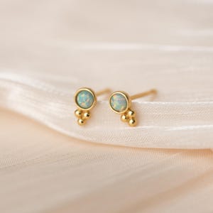 Könnte beinhalten: Ein Paar goldfarbene Ohrstecker. Jeder Ohrring hat einen runden, hellblauen Opalstein, der in einer goldenen Lünette gefasst ist, mit drei kleineren goldenen Perlen, die darunter angeordnet sind. Die Ohrringe werden auf einem weichen, cremefarbenen Stoff präsentiert.