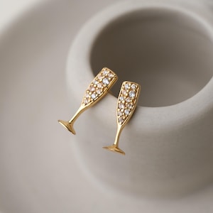 May include: A pair of gold-coloured champagne flute earrings. Each earring is shaped like a champagne flute and is encrusted with small, sparkling gemstones. The earrings are set against a neutral background.