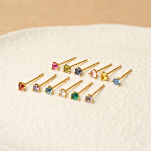 May include: A collection of eleven gold stud earrings, each featuring a single round-cut gemstone in various colors including red, purple, blue, green, yellow, and clear, arranged in a scattered pattern on a textured white surface.