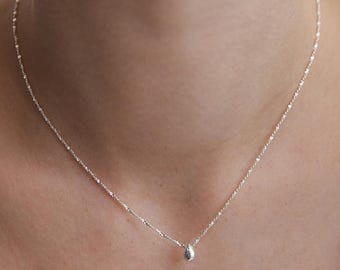 Puede incluir: Un delicado collar de tono plateado que presenta una cadena fina y texturizada con un pequeño dije liso en forma de gota. El dije cuelga centralmente de la cadena que descansa sobre el cuello, mostrando un acabado metálico simple y reflectante.