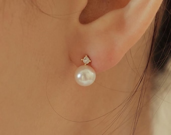 May include: A gold stud earring featuring a small, round, clear gemstone set in a four-prong mount, positioned directly above a smooth, spherical white pearl. The earring is secured to the earlobe, showcasing a classic design with metallic gold accents.