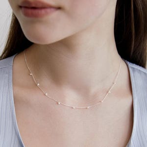 Könnte beinhalten: Eine zarte Silberkette mit kleinen, gleichmäßig verteilten Perlen. Die Halskette liegt am Hals an, wobei die Perlen dezent das Licht reflektieren. Das Design ist schlicht und elegant, geeignet für den Alltag oder besondere Anlässe.