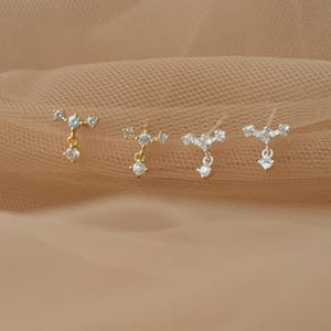 Op de afbeelding: Vier oorknopjes op een rij, elk met een horizontale balk bezet met drie kleine heldere stenen en een enkele bungelende heldere steen. De twee oorbellen aan de linkerkant zijn goudkleurig, terwijl de twee aan de rechterkant zilverkleurig zijn.