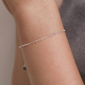 May include: A delicate silver chain bracelet featuring small, evenly spaced metallic beads along the length of the chain. The bracelet has a thin, minimalist profile and includes a small, rectangular silver charm hanging near the clasp area.