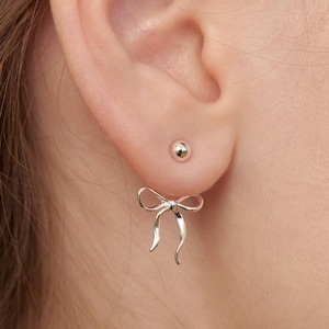 Könnte beinhalten: Nahaufnahme eines silbernen Ohrrings in Schleifenform, der an einem Ohr getragen wird. Der Ohrring hat ein zartes, geschwungenes Design, wobei die Schlaufen und Enden der Schleife deutlich sichtbar sind. Ein kleiner silberner Ohrstecker ist ebenfalls sichtbar.