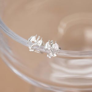 May include: A pair of silver dragon stud earrings. Each earring features a detailed dragon design with outstretched wings and a small, clear gemstone. The earrings are set against a soft, neutral background.
