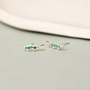 Könnte beinhalten: Ein Paar Ohrstecker aus Silber in Krokodilform, die jeweils mit drei kleinen, runden, leuchtend grünen Steinen entlang des Körpers besetzt sind. Die Ohrringe haben eine polierte metallische Oberfläche und einen Steckerverschluss.
