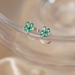 May include: A pair of silver stud earrings, each featuring a four-leaf clover design. The clovers are composed of vibrant green gemstones, adding a pop of color. The earrings are set against a soft, blurred background.
