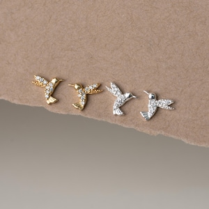 Puede incluir: Cuatro pendientes de botón con forma de colibrí dispuestos en fila. Dos pendientes son de tono dorado con piedras transparentes incrustadas en las alas y el cuerpo, y dos son de tono plateado con piedras transparentes, todos con cierre de presión.