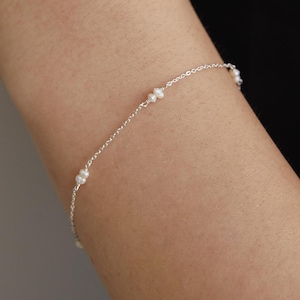 May include: Delicate silver bracelet featuring small, white pearls. The bracelet is a fine chain design with evenly spaced pearls, creating a minimalist and elegant look. The jewelry is shown against a dark background.