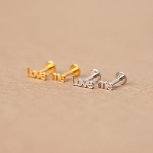 May include: Four small, stud earrings with the words "Love me" in a script font. Two are gold-colored, and two are silver-colored. The earrings have a flat back and a post for piercing. The background is a solid, neutral tan color.