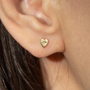 Può includere: Un orecchino a lobo a forma di cuore dorato con una piccola gemma trasparente al centro. L'orecchino è indossato su un orecchio, mettendo in risalto il suo design delicato e la sua sottile lucentezza. È un gioiello semplice ma elegante.