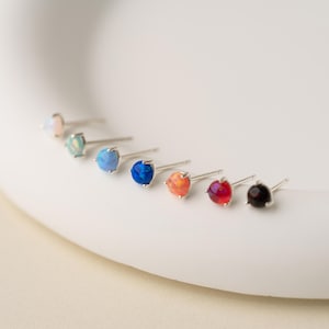 May include: A selection of stud earrings featuring round gemstones in various colours. The earrings are displayed in a row, with colours including pink, teal, blue, orange, red, and black. Each earring has a silver post and setting.