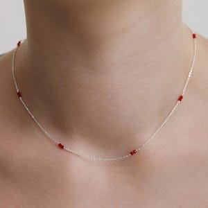 Puede incluir: Un delicado collar de plata con pequeñas cuentas rojas vibrantes espaciadas a lo largo de la cadena. El collar se muestra sobre un fondo neutro, destacando el diseño minimalista y el contraste de la plata y el rojo.