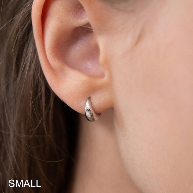 May include: A small, silver hoop earring is worn on an ear. The earring is a simple, rounded design, reflecting light. The word "SMALL" is printed in white at the bottom of the image.