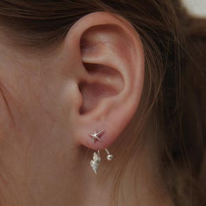 May include: A silver earring featuring a small starfish stud on the front of the earlobe, with a dangling seashell charm and a spherical bead attached to the back post, creating a layered look that sits below the bottom edge of the earlobe.