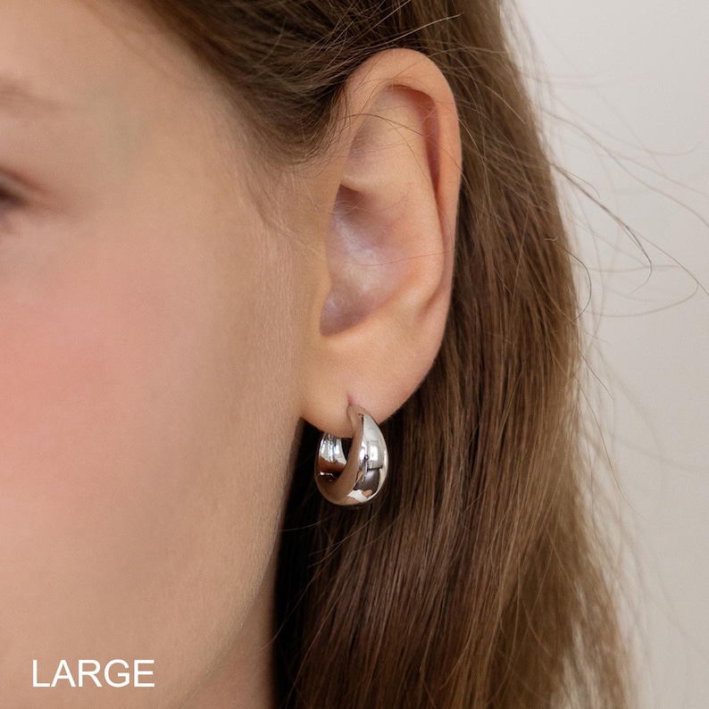 May include: A close-up of a silver hoop earring. The earring is a thick, rounded design, reflecting light. The word "LARGE" is printed in white at the bottom of the image. The earring is worn on a person's ear.