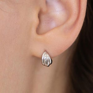 Puede incluir: Un pequeño pendiente de aro plateado con una textura acanalada similar a una concha. El pendiente presenta un acabado pulido y reflectante, con una forma semicircular curva que se ajusta al lóbulo de la oreja y un diseño de tres secciones.