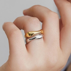May include: Two smooth, organic-shaped rings. One ring is gold-colored, and the other is silver-colored. The rings are stacked on a finger, showcasing their polished, reflective surfaces. The jewelry is simple and elegant.