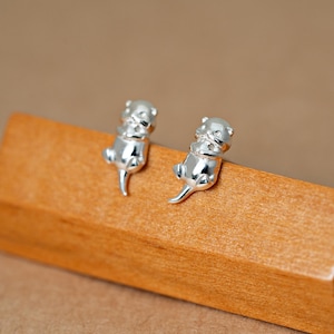 Peut inclure: Une paire de boucles d'oreilles à clous argentées en forme de petites loutres. Chaque boucle présente une finition métallique polie avec un corps lisse et arrondi, une queue pointue et des traits du visage détaillés, représentant l'animal couché.