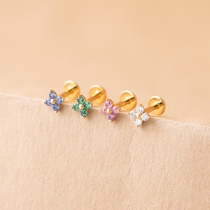 May include: Four gold-colored flower-shaped earrings with colorful gemstones. The gemstones are blue, green, pink, and white. The earrings are displayed on a light brown surface.