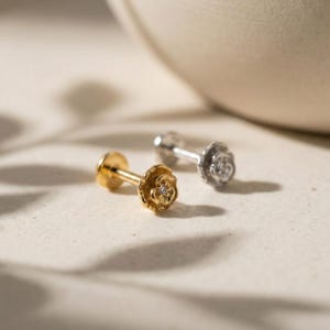 Pode incluir: Dois brincos pequenos com um design floral. Um é dourado e o outro prateado. Cada brinco tem uma pequena pedra preciosa redonda no centro da flor. Os brincos estão sobre uma superfície clara.